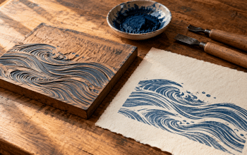 Woodblock carving tools, blue pigment, and an original wave-pattern print on a warmly lit wooden worktable.