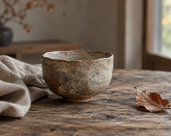 An irregular ceramic tea bowl beside rumpled linen and a fallen autumn leaf on a weathered wooden table in soft window light
