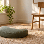 A sage crescent meditation cushion on a woven mat beside a wooden chair and a sunlit window.