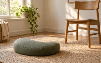 A sage crescent meditation cushion on a woven mat beside a wooden chair and a sunlit window.